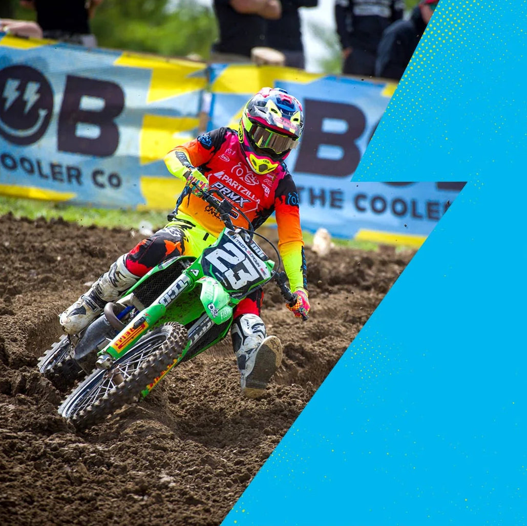 A motocross rider in a bright orange, yellow, and blue racing suit leans sharply into a turn on a green dirt bike over brown dirt track, with a blue and yellow sponsor banner in the background.
